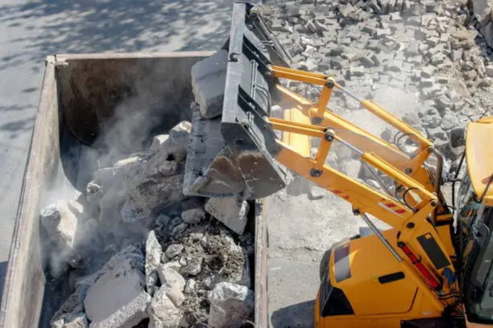 A yellow excavator is actively dumping rubble into a dumpster at a construction site in San Jose, California.